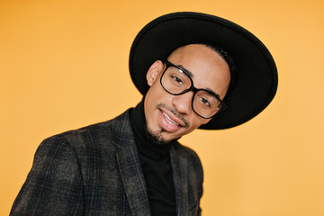 Close-up portrait of interested african guy in trendy glasses. Indoor photo of enchanting black man smiling on orange background.