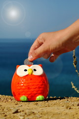 Owl piggy bank on the seashore and hand with a coin. How to save on vacation?