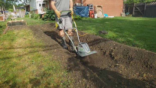 Home Gardening - Man in shorts wearing boots use corded electric tiller or cultivator machine to create new garden bed on his back yard.