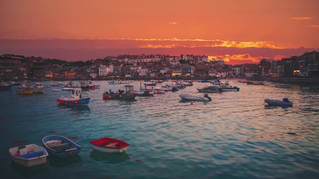 Cinemagraph / seamless video loop of the beach and harbor promenade of the famous town St. Ives in Cornwall in Southern England, UK with fishing boats moving in the clear blue water by sunset.