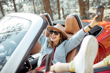 Young stylish woman enjoying traveling by convertible car in nature, sitting freely with raised hands on the driver's seat. Carefree lifestyle and travel concept