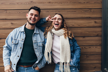 Dreamy girl wears denim jacket and knitted scarf chilling with boyfriend. Indoor photo of amazing female model posing on wooden background with brunette man.