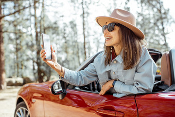 Woman traveling by car in the forest, leaning out of the car window and photographing with a smart...
