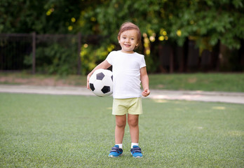 Little soccer player: toddler boy in sports uniform standing on green stadium holding football ball...