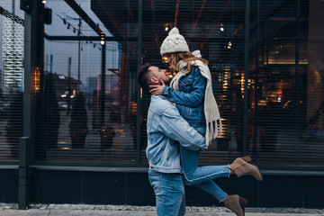 Graceful girl in knitted scarf and high heel boots having fun during date. Outdoor portrait of european guy holding his girlfriend on urban background.