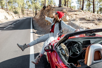 Lifestyle portrait of a carefree woman dressed casually in jeans and red hat sitting on the car hood, enjoying road trip on the mountain road