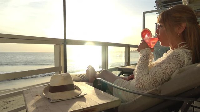 Woman relaxing with a refreshing drink on a scenic oceanfront deck overlooking the beach at sunset