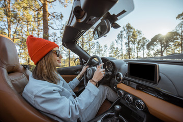 Happy woman dressed casually in jacket and red hat driving convertible car on the beautiful...