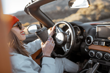Happy woman driving convertible car with coffee cup while traveling on the desert road. Carefree...