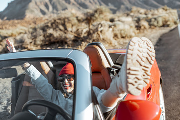 Woman traveling by convertible car on the desert valley, pulling legs out of the car window on the roadside. Lifestyle in travel concept