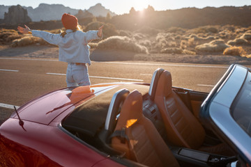 Woman enjoying beautiful rocky landscapes while standing with a car on the desert roadside during a sunset. Traveling on the volcanic valley on Tenerife island