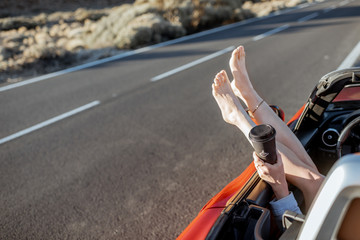 Woman pulling legs out of the car window on the roadside. feeling comfortable while traveling. Close-up on female legs and coffee cup