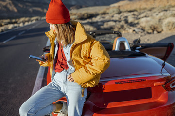 Stylish woman in yellow jacket and red hat enjoying roadtrip, sitting with a smart phone on the car...