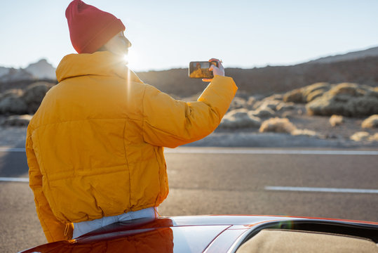 Woman enjoying beautiful volcanic landscapes, photographing on phone on the roadside during a sunset. Traveling on the desert valley on Tenerife island