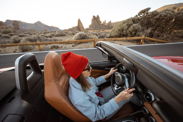 Happy woman in red hat driving convertible car while traveling on the desert road with beautiful...