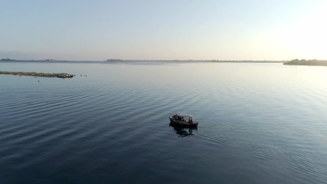 Aerial view of a boat sailing along the deep blue waters of the Ambracian Gulf in Greece  going towards an aquaculture with the sun reflecting in the back, drone footage