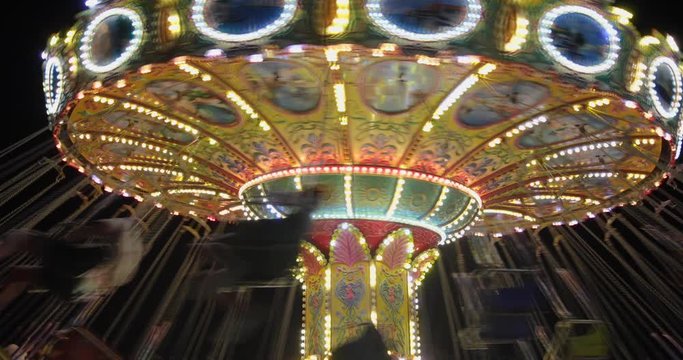 A fast spinning merry go round or carousel with seats suspended from the top at night. It is beautifully decorated with colored flashing lights. People are flying in a circle