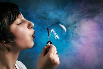 Fototapeta premium Portrait of a teenager girl blowing soap bubbles on an abstract background with multi-colored texture smoke.