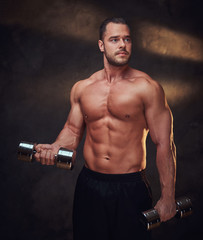 Fototapeta na wymiar Attractive muscular man is doing exercises with dumbbells at dark room.