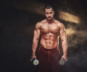 Fototapeta na wymiar Young handsome bodybuilder is posing for photographer with dumbbells.