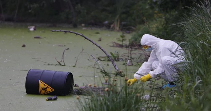 A scientist in a protective suit takes a water sample from a polluted lake next to a black barrel with a bio-hazard sign.
