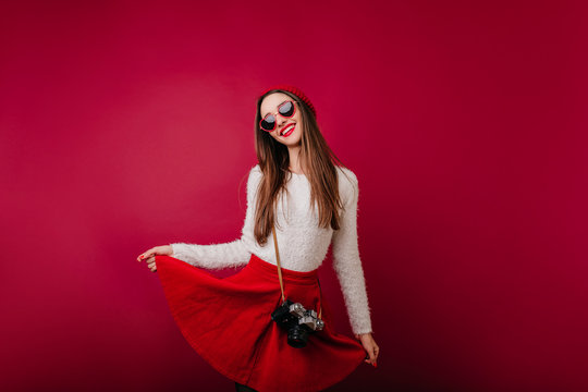 Fashionable smiling girl playing with her red skirt. Studio shot of brunette woman with camera enjoying photoshoot.