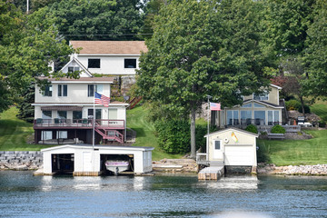 Different buildings in a grassy area on the coast of a lake during daytime, and surrounded by trees. United States flags, and boat cottage. Real State concept. Thousands Islands. Ontario, Canada/USA