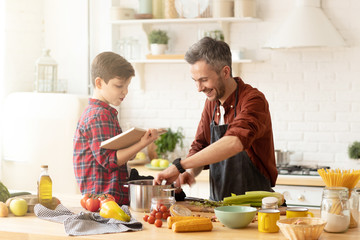 Son reading cookbook to dad cooking dinner at modern bright loft kitchen.