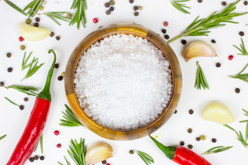 Top view of white sea salt in a round wooden bowl with flat lay red chilli pepper, peppercorns, garlic and rosemary on food background.