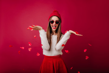Graceful girl in fluffy sweater throwing out heart confetti. Lovable female model in red skirt...