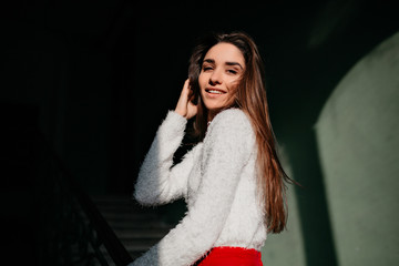 Caucasian woman with straight hair posing near stairs and smiling. Positive young lady in fluffy sweater standing in the dark.