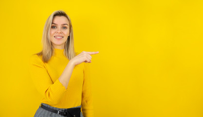 Amazing young woman points finger the side. Caucasian model smiling and looking in camera. Isolated on yellow background. Copy space