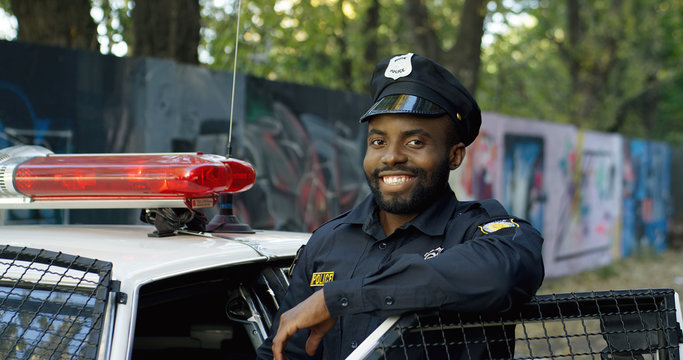 Portrait of handsome African American young policeman in uniform and cap. Front view of police officer smiling at camera outdoors. Cop on the background with police car and old lane.