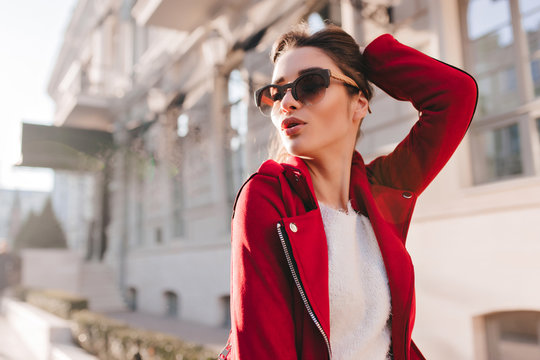 Sensual woman in big sunglasses playing with her hair in sunny day. Outdoor photo of relaxed caucasian girl wears red jacket.