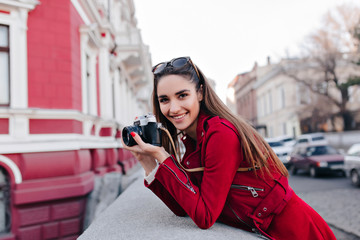 Lovely white woman in stylish red attire taking picture of street view. Elegant funny photographer smiling during outdoor photoshoot.