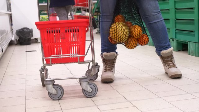 New environmentally friendly packaging for supermarket products. Mesh knitted from natural cotton threads. Adult girl put oranges in net and waving it next to grocery cart