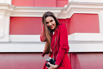 Graceful female photographer with happy face expression playing with her hair. Refined caucasian girl in red clothes standing on the street with camera.