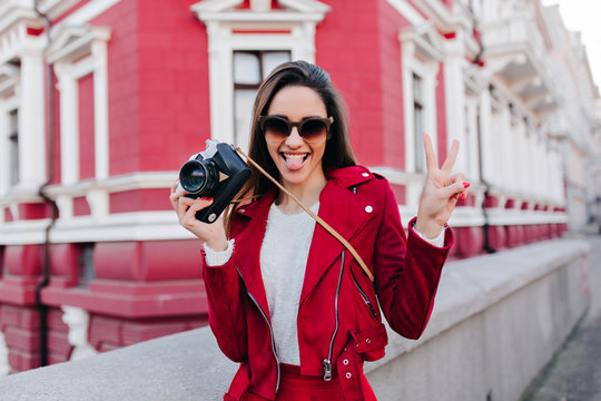 Ecstatic brunette girl posing with peace sign and tongue out. Spectacular young woman in red outfit standing with camera and laughing on house background.