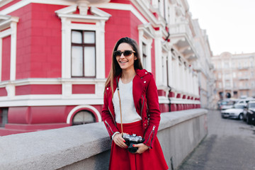 Lovable slim girl with camera expressing happiness. Outdoor photo of enthusiastic female photographer wears stylish sunglasses.