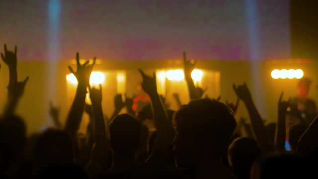 Slow motion: people crowd partying, raising hands up, showing sign - devil's horns gesture and jumping at rock concert in front of stage of nightclub. Nightlife and entertainment concept