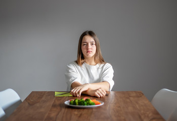 Teen girl sitting at table with crossed hands and unhappy face in front of plate with broccoli and...
