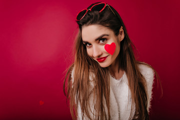 Close-up portrait of playful girl in trendy sunglasses isolated on claret background. Studio shot of brunette refined woman with heart on cheek.