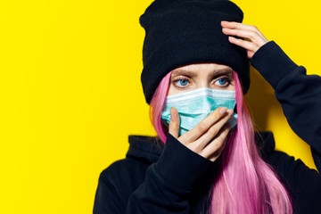 Studio portrait of young girl with pink hair and blue eyes, wearing medical flu mask, dressed in black hoodie sweater and beanie hat on yellow background.