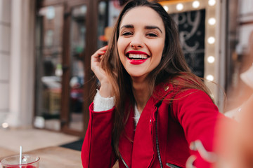 Close-up portrait of girl with gorgeous smile making selfie in red outfit. Outdoor photo of positive dark-haired lady taking picture of herself and expressing happiness.