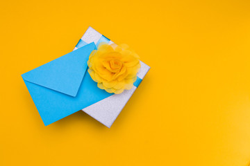 a blue envelope lies under a silver shiny box with a blue ribbon and a large yellow flower of light delicate fabric, yellow background, copy space, top view