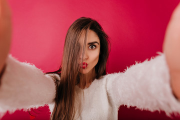 Surprised girl with big eyes making selfie on claret background. Gorgeous young lady in white...