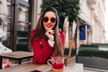 Positive dark-haired girl sitting in cafe near green tree. Outdoor portrait of glamorous young woman in red enjoying tea on city background.