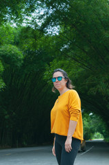 Woman with yellow sweater and sunglasses, with direct look, on bamboo path, open view, bus stop