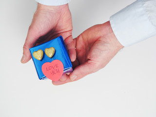 Male holding blue gift box with two golden hearts and a red heart shaped sticker with sign love you. Love and romance theme. White background.