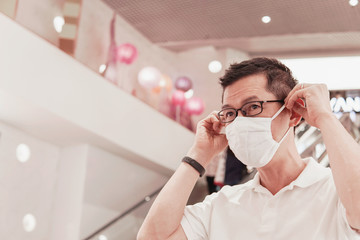 Middle aged Asian man wearing glasses and medical face mask in shopping mall,  covid-19 coronavirus...